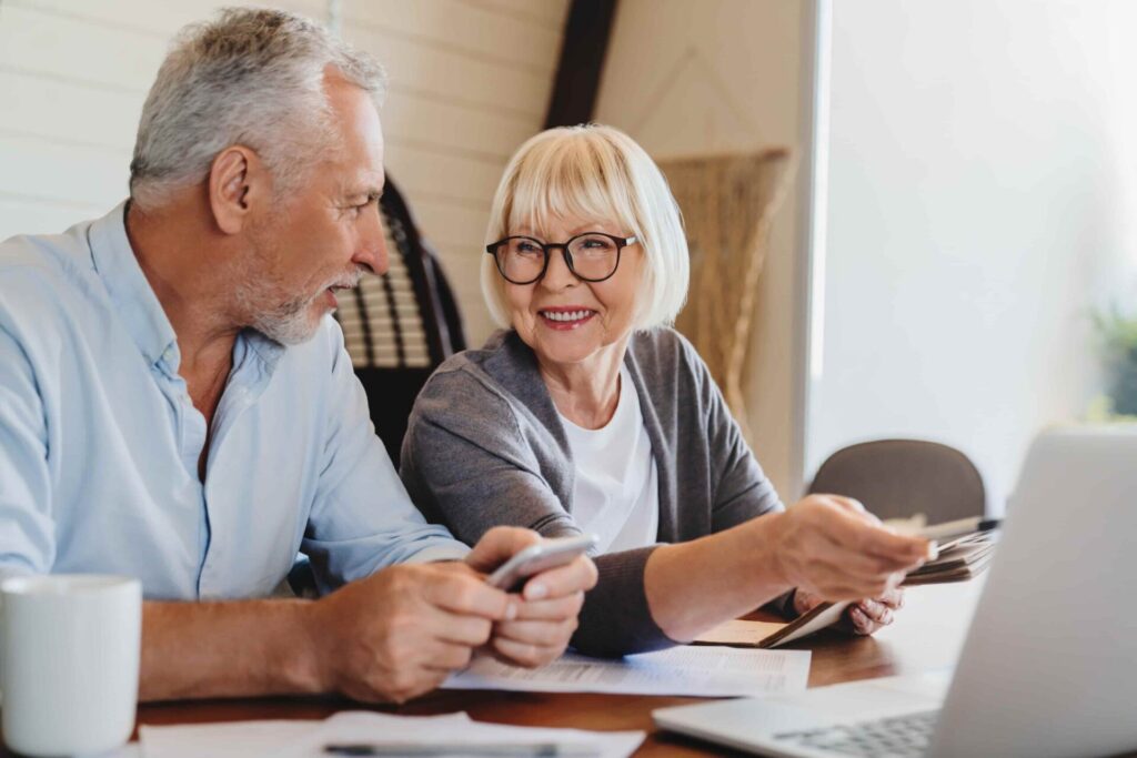 Cheerful older couple sitting at table with computer and paperwork, assessing asset location strategy