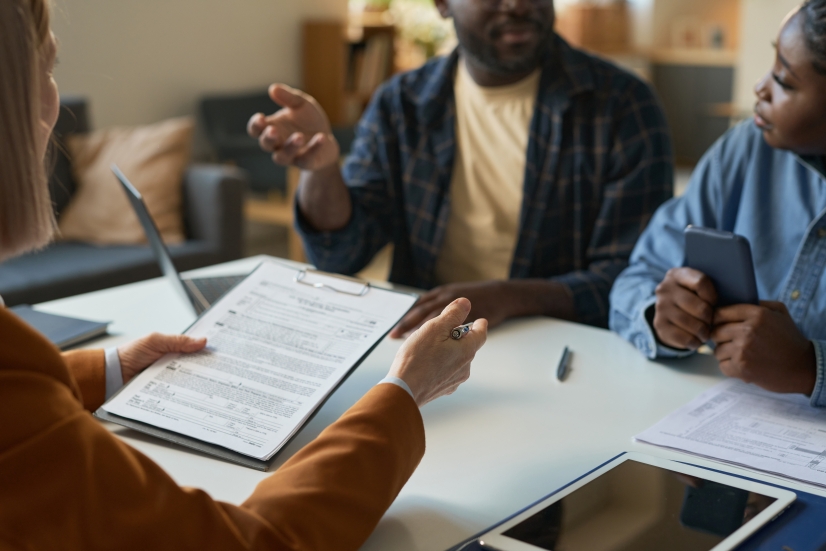Close up of couple sitting at a table across from a professional holding documents on a clipboard, discussing how to avoid tax mistakes.